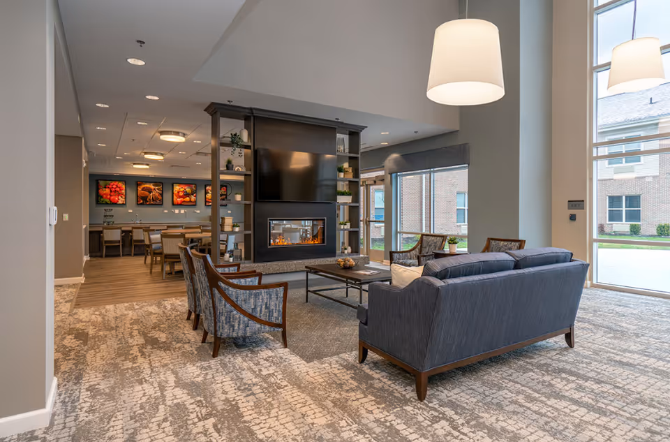 A modern senior living common area with a seating arrangement including a blue sofa and patterned armchairs around a coffee table. A large fireplace with a TV mounted above it is centrally located, and there are large windows letting in natural light. In the background, there is a dining area with tables and chairs, and colorful food-themed artwork on the wall.