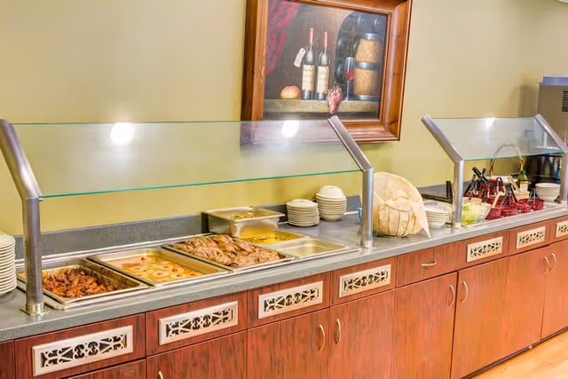 Buffet counter with a glass sneeze guard displaying trays of food including chicken wings, dessert with cherries, baked goods, and corn. Plates and bowls are stacked at the end of the counter. A framed painting of wine bottles and fruit hangs on the wall above the buffet.