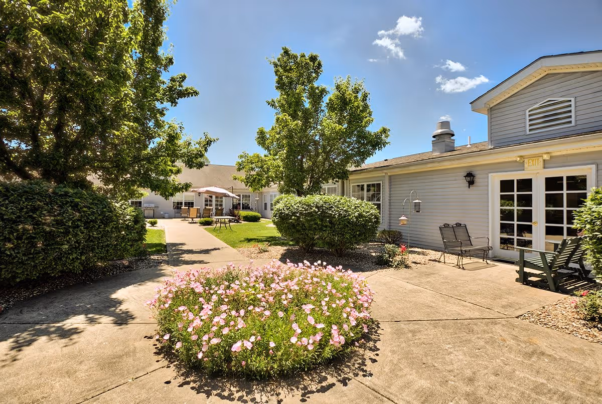Outdoor courtyard area at Cardinal Place featuring a circular flower bed with pink flowers in the center, surrounded by concrete walkways. There are green bushes and trees providing shade, and patio furniture including chairs and tables with umbrellas near the building. The building has light gray siding and white-framed windows and doors under a clear blue sky.