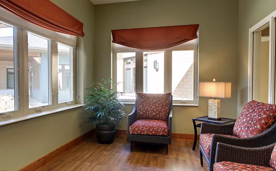 A cozy sitting area with two cushioned armchairs and a small side table with a lamp, positioned near large windows with red valances. There is a potted plant in the corner and wooden flooring.