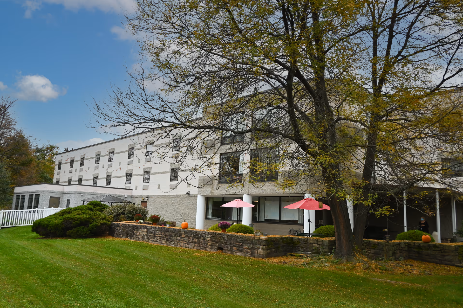 Exterior view of a multi-story senior living facility building with a stone retaining wall, green lawn, and trees with autumn foliage. There are two pink umbrellas on a patio area near the building entrance.