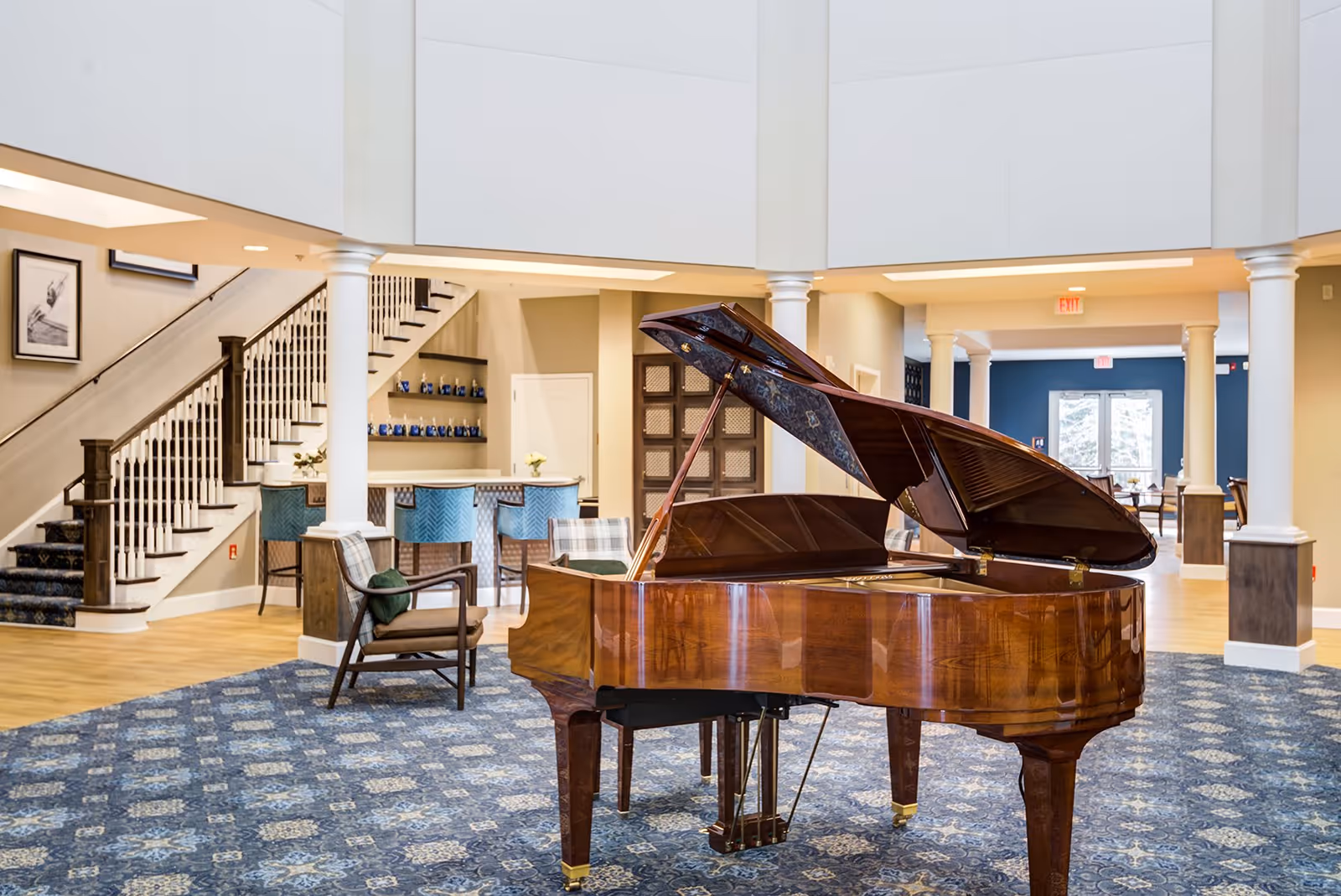 Spacious senior living facility common area with a polished grand piano in the center on a patterned blue carpet. The room features white columns, a staircase with dark wood railing and carpeted steps, a seating area with chairs, and a bar counter with blue stools. The walls are painted in neutral tones with some blue accents, and there are framed pictures on the wall near the staircase.