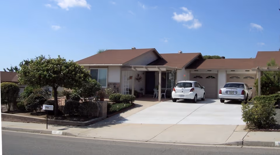Single-story residential building with a brown roof and white exterior walls. There are two cars parked in the driveway, a white compact car and a white sedan. The front yard has neatly trimmed bushes, a small tree, and a mailbox near the street. The sky is clear with a few clouds.
