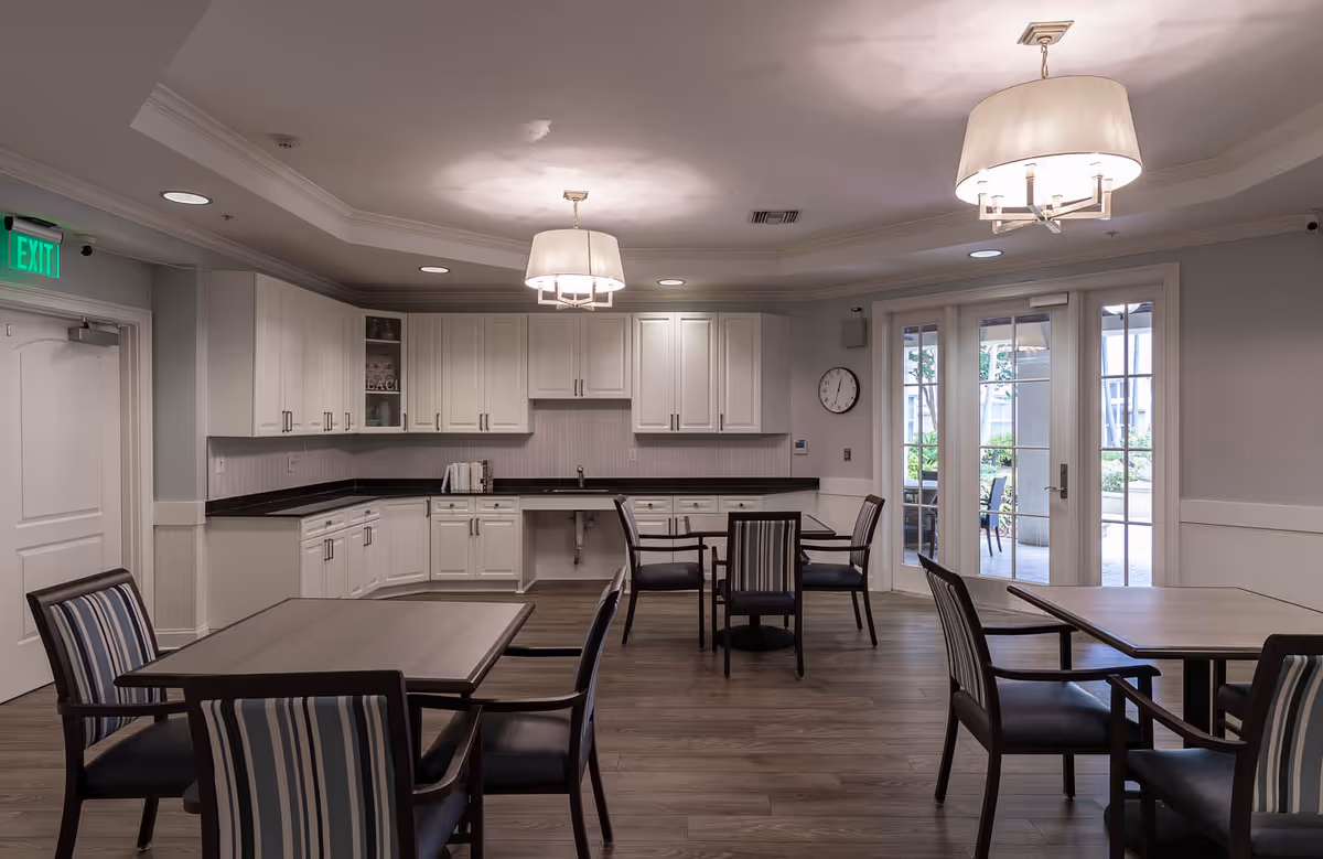 Interior view of a communal dining area with several tables and chairs. The room features a kitchenette with white cabinets and a black countertop along one wall. Two modern pendant lights hang from the ceiling, and there is a clock on the wall near glass doors that lead to an outdoor patio area.