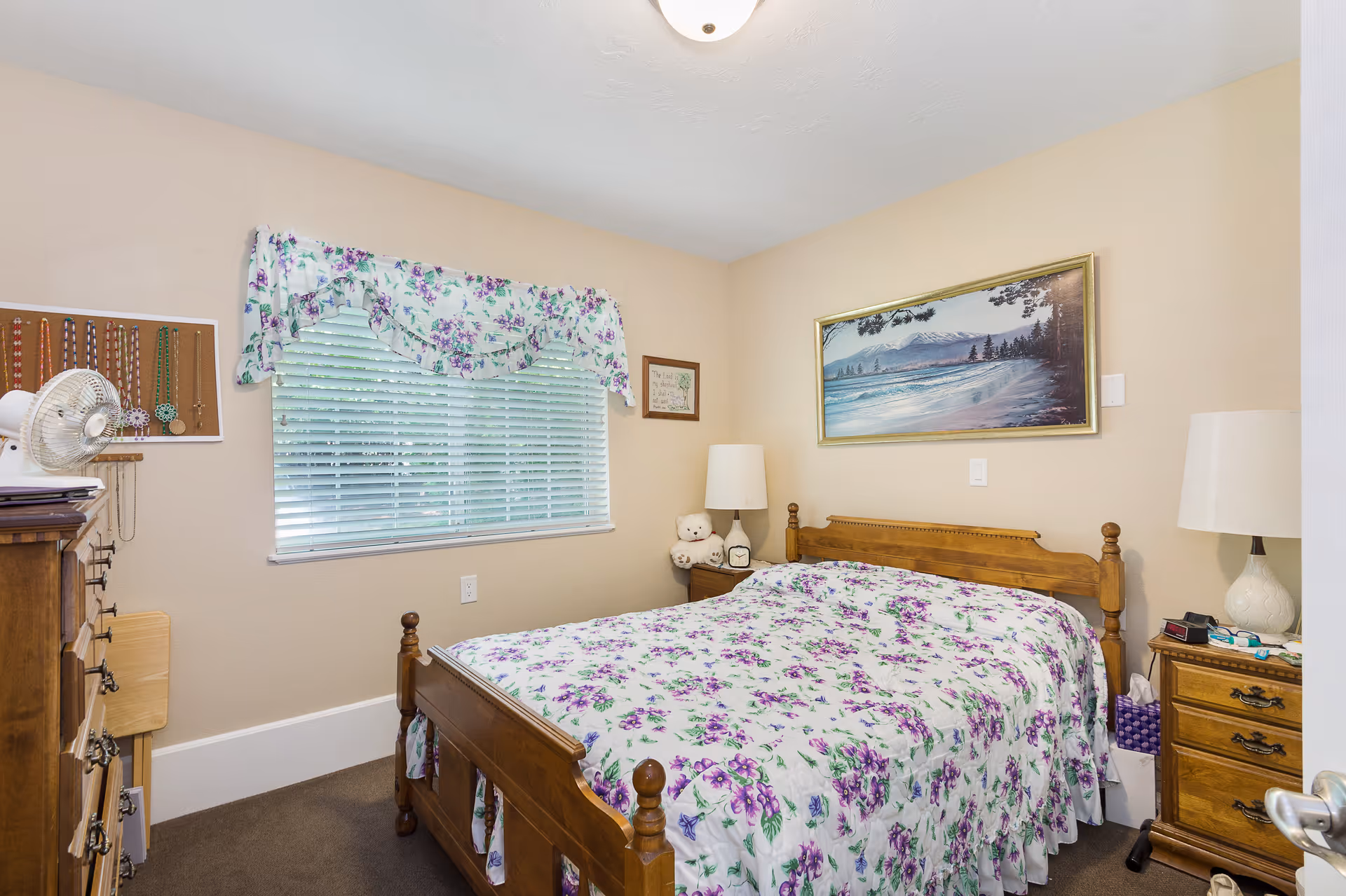 A cozy bedroom with a wooden bed frame and matching furniture. The bed is covered with a white bedspread featuring a purple floral pattern. A window with white blinds and a floral valance is on the left wall. There are two bedside tables with lamps, a teddy bear, and other small items. A landscape painting hangs above the bed on the beige wall.