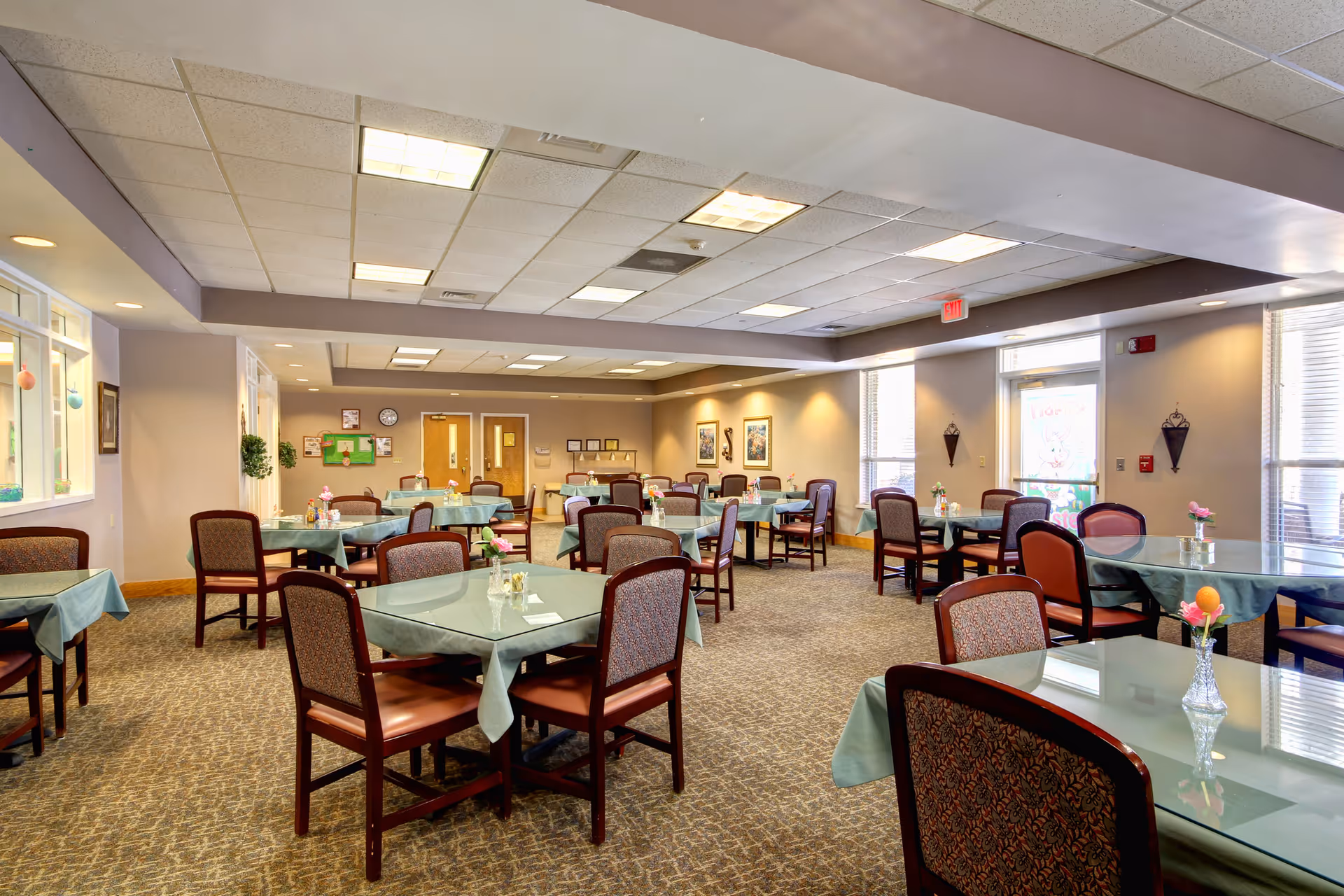 Dining room with multiple tables covered in teal tablecloths, upholstered chairs, and large windows letting in daylight.