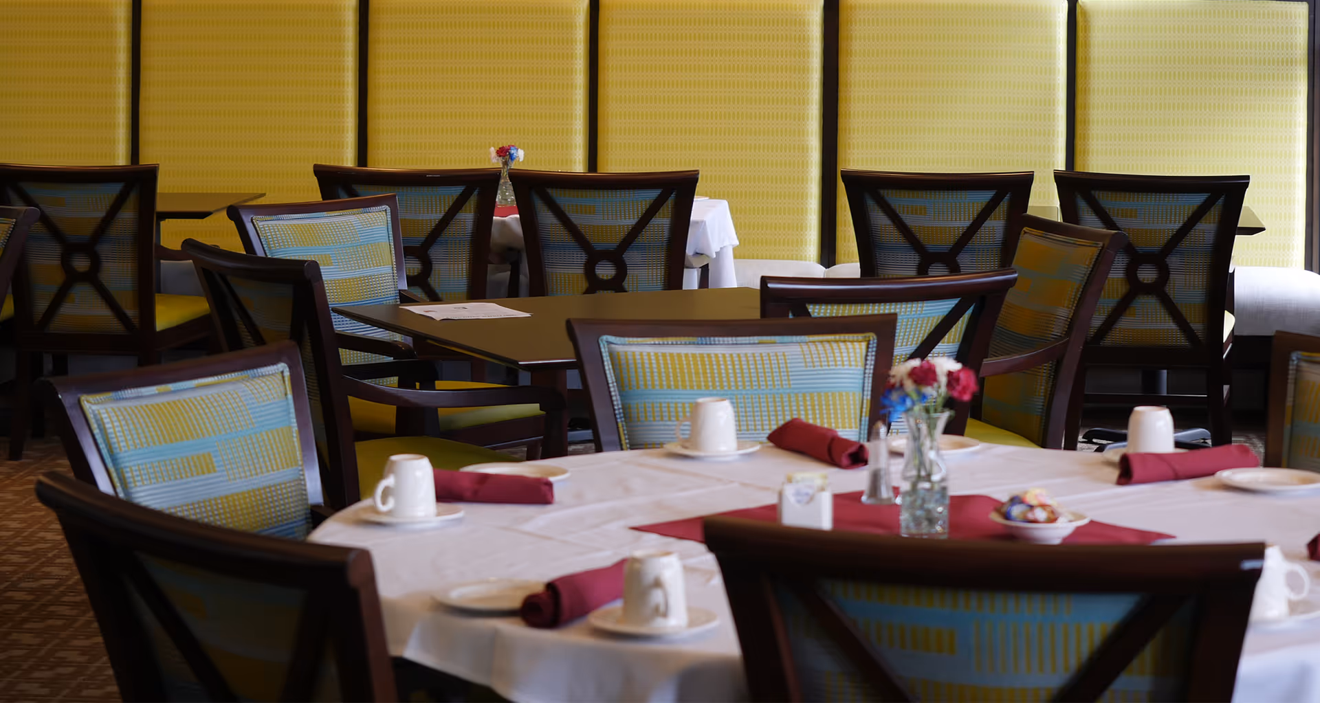 Dining area with tables set for a meal, featuring white tablecloths, maroon napkins, white cups on saucers, small plates, and a vase with flowers on the table. Chairs have wooden frames with patterned upholstery in blue and yellow. The background shows a yellow padded wall.