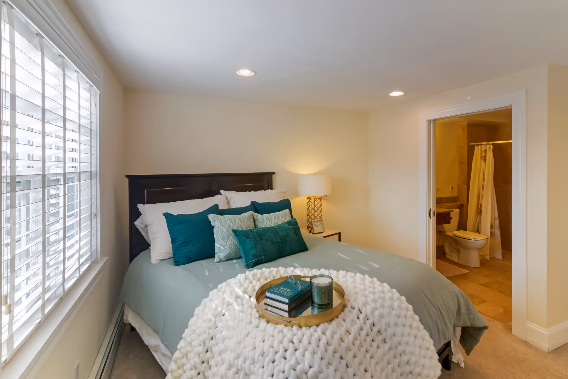 A cozy bedroom with a neatly made bed featuring teal and white pillows, a chunky white knit blanket, and a tray holding books and a candle. There is a window with white blinds on the left side, a bedside table with a lamp and a clock, and an open door leading to a bathroom with a toilet and shower visible.