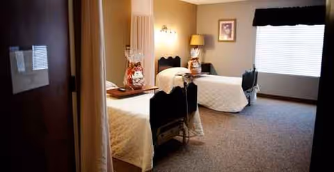Two twin beds in a dimly lit senior living bedroom with nightstands and window.