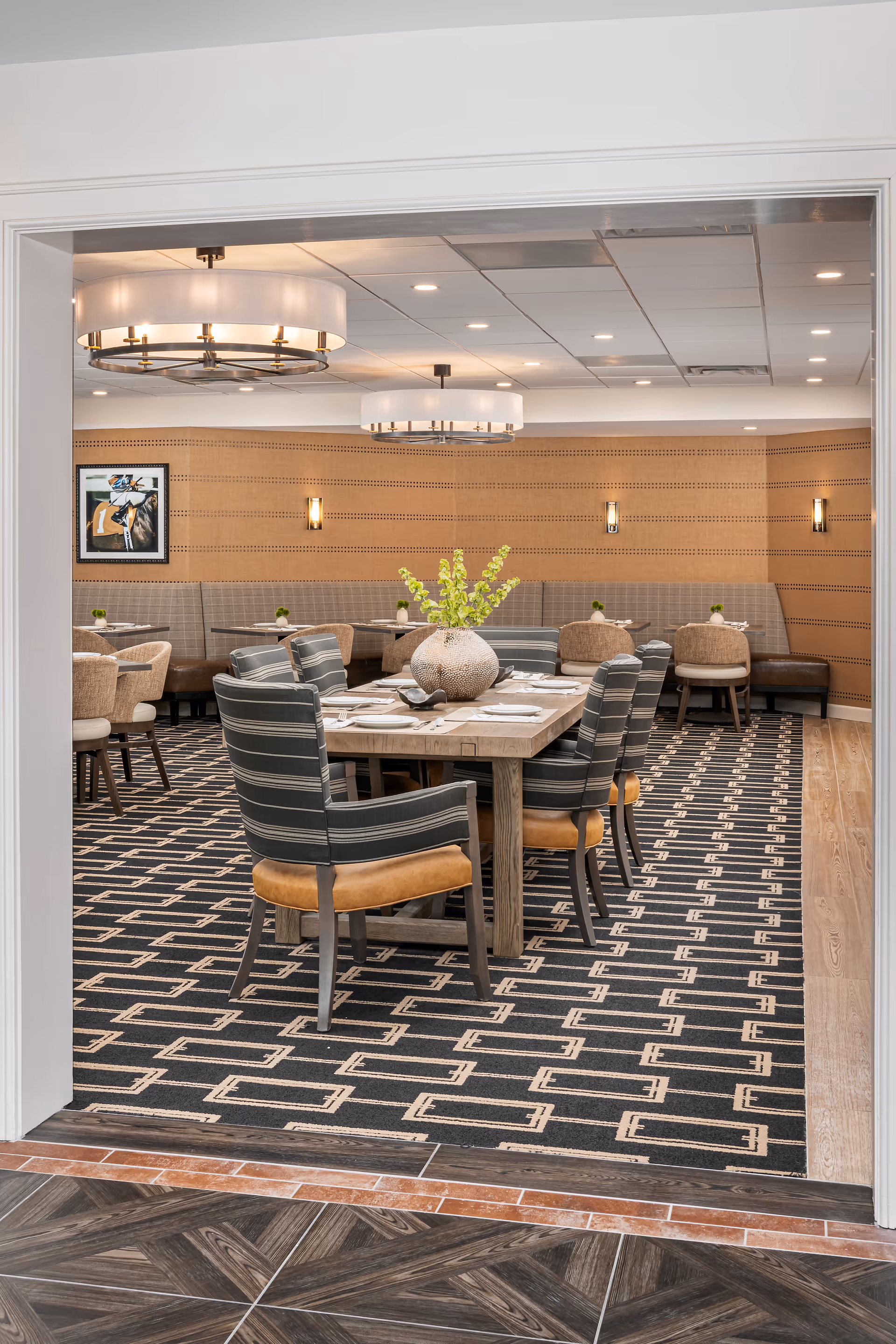 A modern dining room with a rectangular wooden table set for six people. The chairs around the table have striped upholstery and tan leather seats. The room features a patterned carpet, beige walls with horizontal lines, wall sconces, and two large circular ceiling lights. Additional seating with round tables and chairs is visible in the background, along with a framed artwork on the wall.