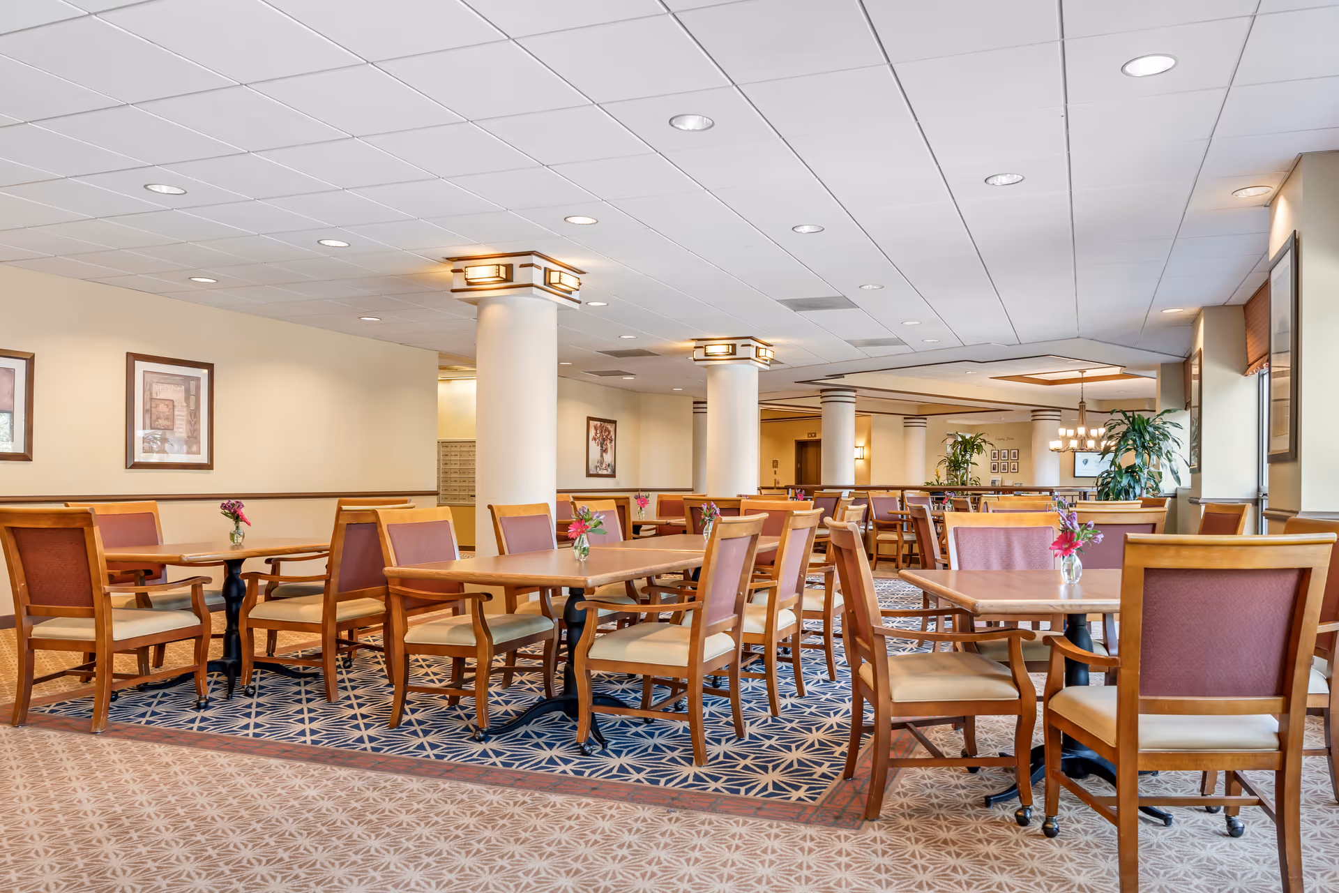 A spacious dining room with multiple wooden tables and chairs arranged neatly. Each table has a small vase with flowers. The room features patterned carpeting, white ceiling tiles with recessed lighting, and several framed artworks on the walls. Large windows on the right side allow natural light to enter, and there are decorative plants placed around the room.