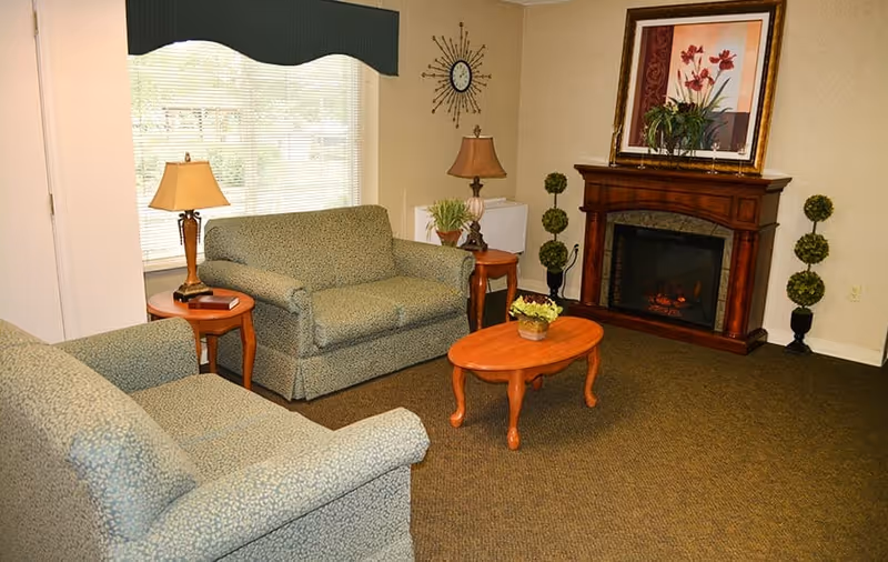 A cozy living room with two patterned armchairs and a loveseat arranged around a wooden coffee table with a small plant. There are two side tables with lamps, a wall clock, a framed floral painting above a wooden electric fireplace, and decorative topiary plants on either side of the fireplace. A window with blinds and a dark valance is visible in the background.