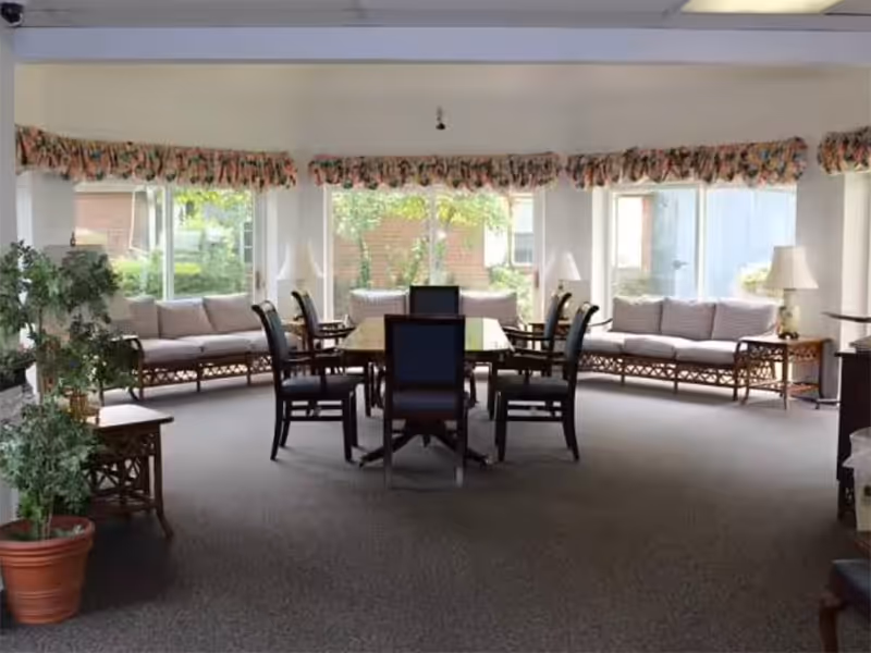 Communal sitting area with a central dining table surrounded by chairs, sofas along large windows, and potted plants.