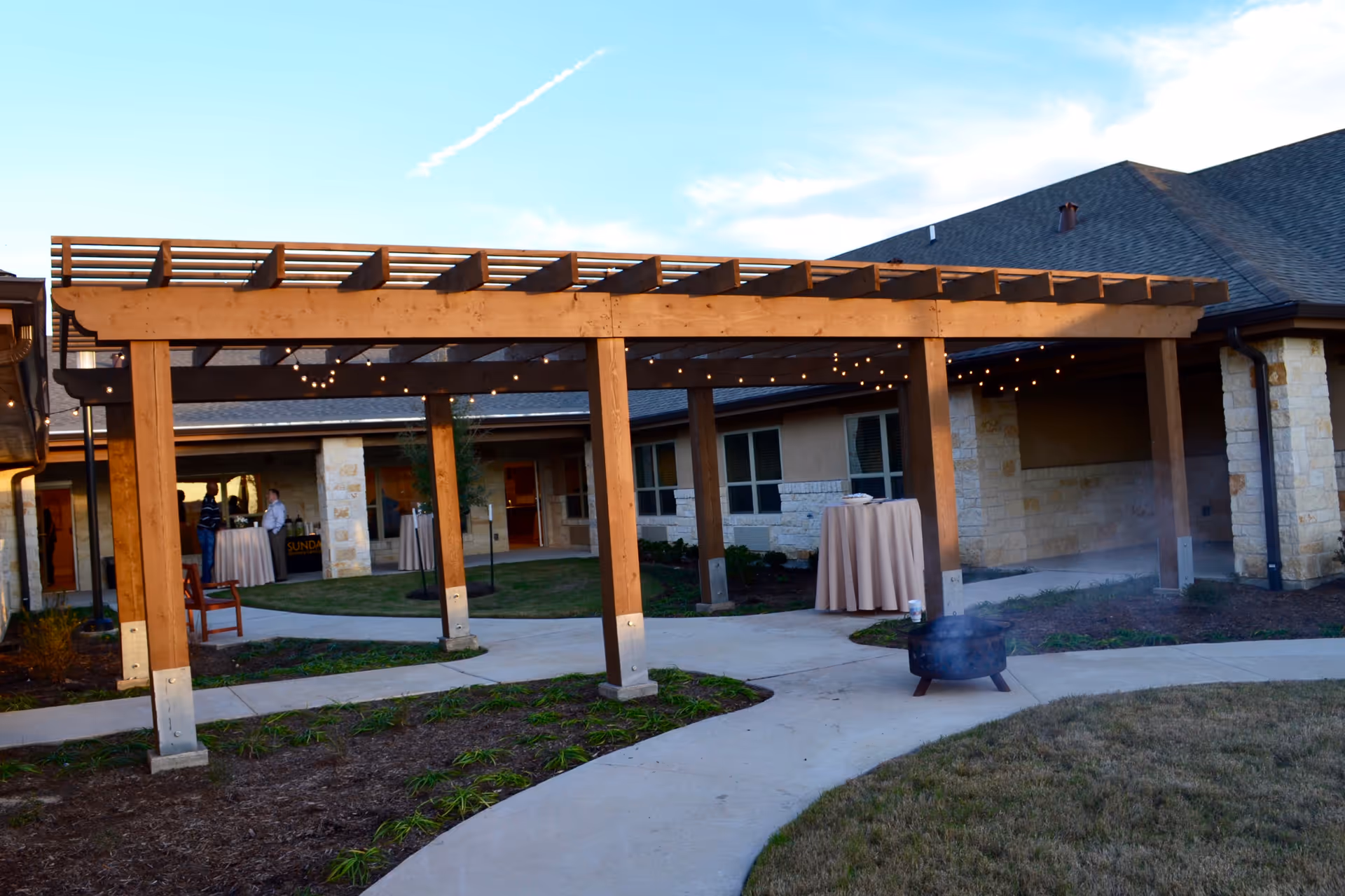 Outdoor patio area at Sundance Memory Care at Towne Lake featuring a wooden pergola with string lights, a curved concrete walkway, a small fire pit emitting smoke, and a few tables covered with beige tablecloths near the building entrance.