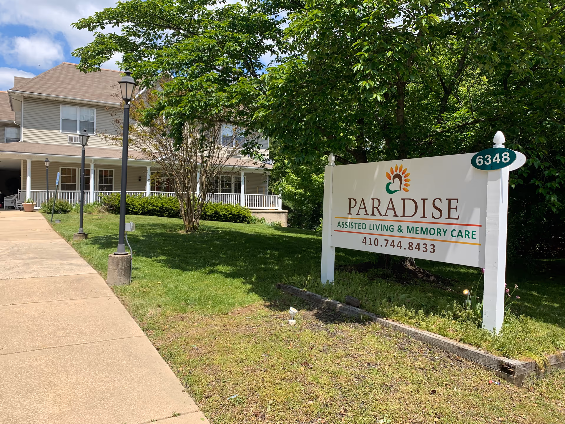 Exterior view of Paradise Assisted Living & Memory Care facility showing a large sign with the facility's name, phone number, and address number 6348. The building has a beige exterior with white trim, a covered porch, and is surrounded by green grass, trees, and lamp posts along a concrete walkway.