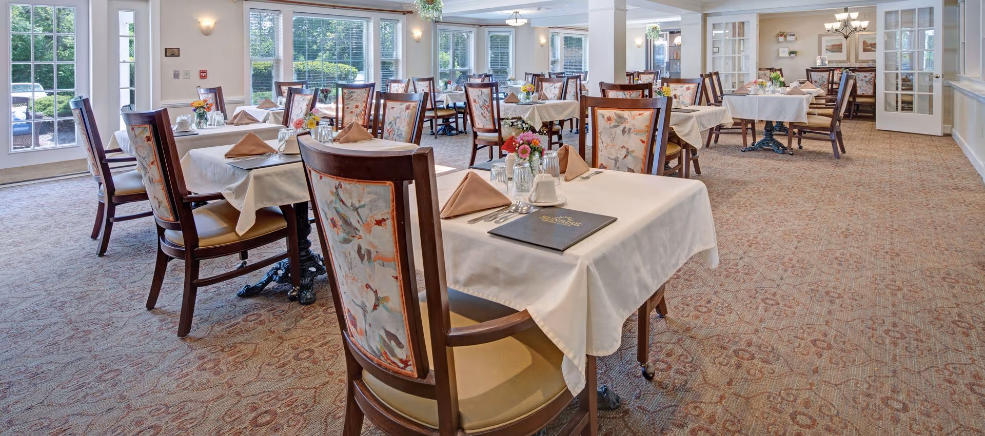 Well-lit dining room with multiple tables set with white tablecloths, floral-backed chairs, and place settings.