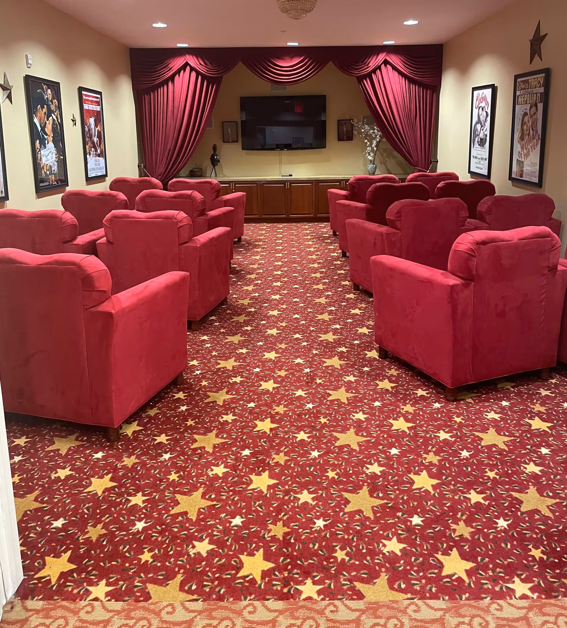 A small theater room with red upholstered armchairs arranged in rows facing a flat screen TV mounted on the wall. The room features red curtains framing the TV, a red carpet with yellow star patterns, and movie posters on the walls.