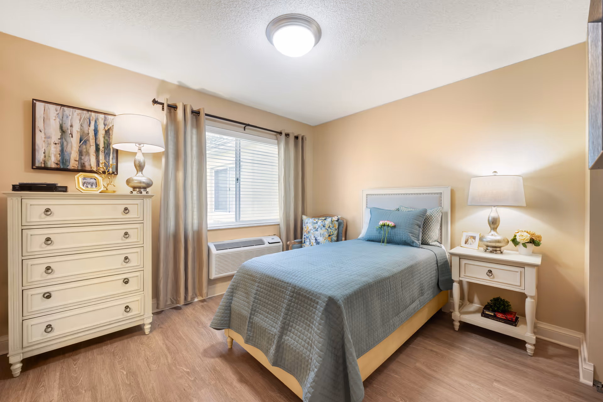 A cozy bedroom with a single bed covered in a blue quilt and matching pillows. There is a white nightstand with a lamp, a small framed photo, and a flower pot on it. Next to the bed is a cushioned chair with a floral pattern. On the opposite side of the room is a white chest of drawers with a lamp, a framed photo, and a decorative item on top. A window with beige curtains and a wall-mounted air conditioning unit is centered on the wall. The room has light-colored walls and wood flooring.