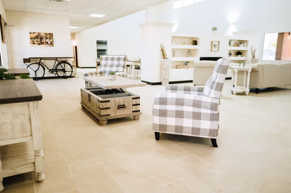 A bright and spacious living room area with beige tiled flooring, two checkered armchairs, a wooden coffee table with a lift-top, beige sofas, built-in white shelves with decorative items, and a bicycle-shaped console table against the wall.