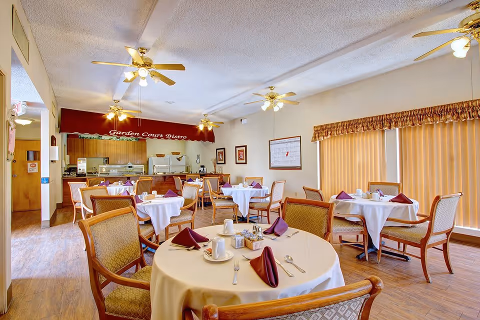 Dining room with round tables covered in white tablecloths, each set with purple napkins, cups, and silverware. Wooden chairs with cushioned seats surround the tables. Ceiling fans with lights hang from the ceiling. In the background, there is a serving area with a red awning labeled 'Garden Court Bistro' and kitchen appliances. Large windows with vertical blinds allow natural light into the room.