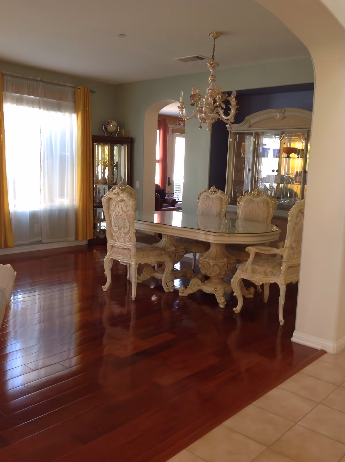 A dining room with a polished wooden floor and a large ornate dining table with six matching upholstered chairs. A chandelier hangs above the table. There is a large window with sheer white curtains and yellow drapes on the left side. In the background, there is a glass display cabinet with decorative items and an arched doorway leading to another room.
