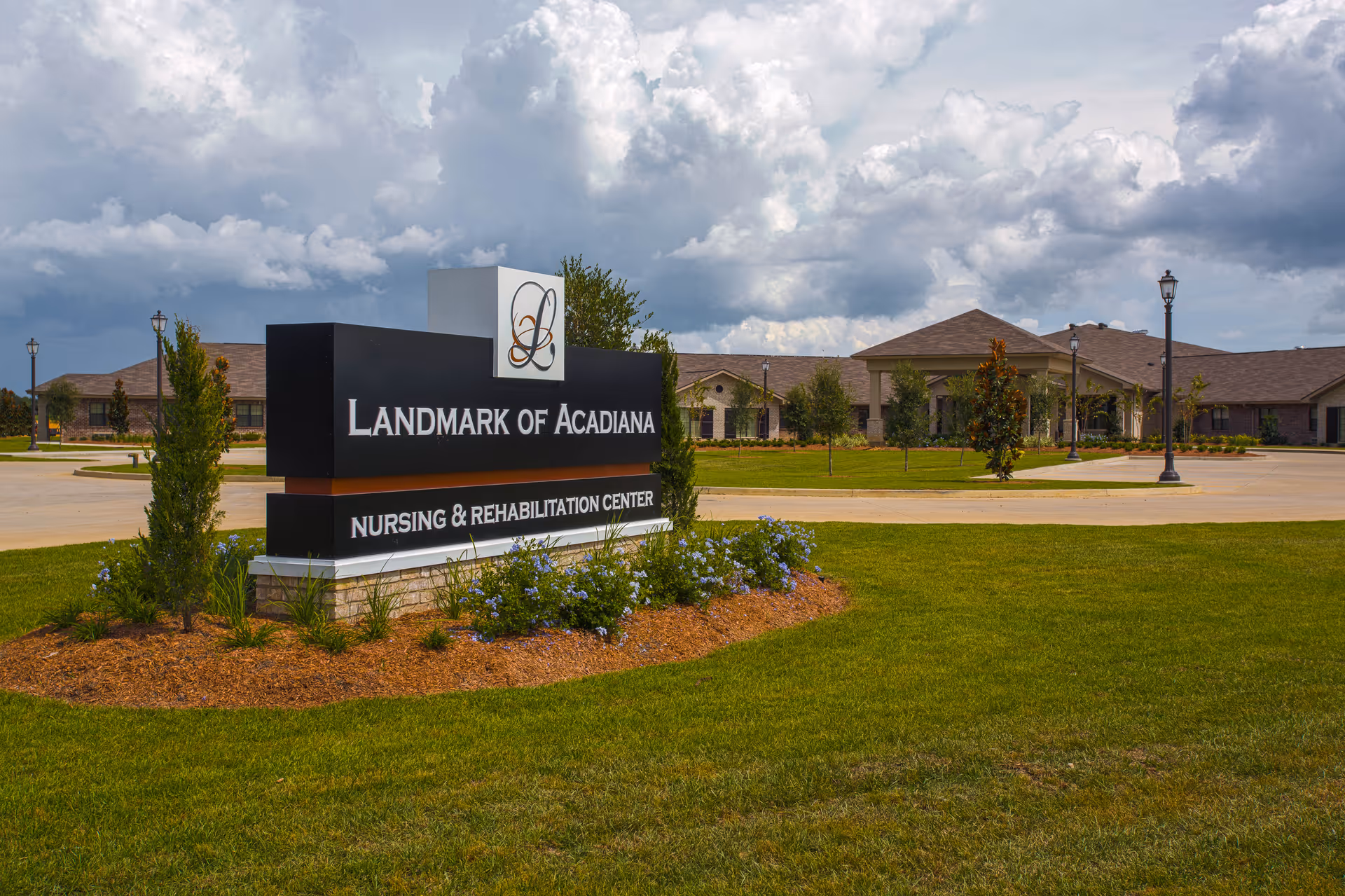 Entrance sign for Landmark of Acadiana nursing and rehabilitation center on a landscaped lawn with the facility building and cloudy sky in the background.