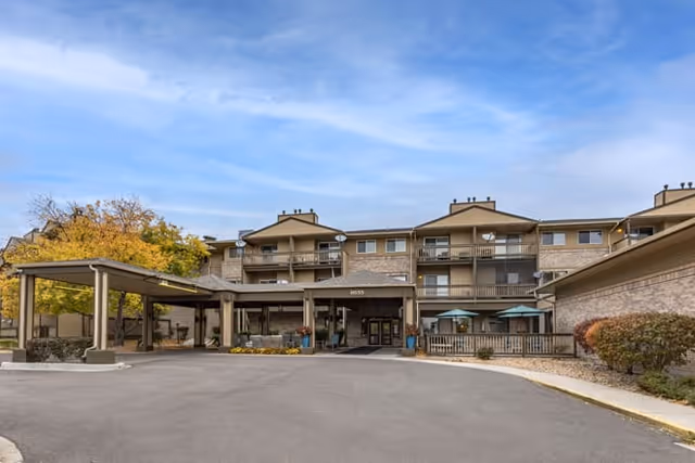 Front exterior view of a multi-story senior living facility building with balconies, a covered entrance, and a driveway. Trees with autumn foliage and a clear blue sky are visible.