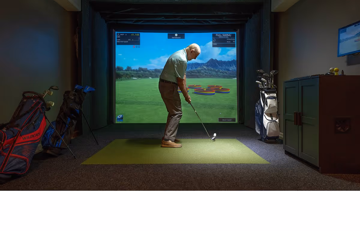 An elderly man playing golf indoors using a golf simulator. He is standing on a green mat with a golf club, aiming at a large screen displaying a virtual golf course. Several golf bags with clubs are placed around the room.