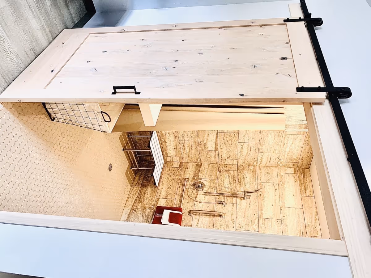 View of a bathroom shower area with beige tiled walls and floor, a metal shower bench, grab bars, a handheld showerhead, and a red and white shower chair. The shower is partially enclosed by a light wood sliding barn door with black hardware.