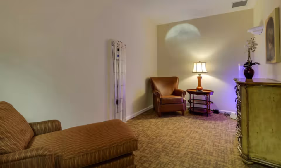A cozy room with a brown upholstered chaise lounge on the left, a brown leather armchair next to a round wooden side table with a lit lamp, and a vintage-style dresser with a vase of flowers on top. The walls are light-colored and the floor is carpeted.