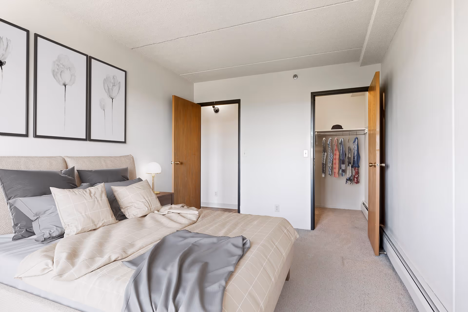A bedroom with a neatly made bed featuring beige and gray bedding. Above the bed are three framed black and white floral prints. To the right, there are two open wooden doors, one leading to a hallway and the other to a closet with clothes hanging inside. The room has light-colored walls and carpeted flooring.