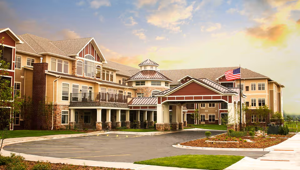 Front exterior of a large multi-story senior living facility with a covered porte-cochere, balconies, landscaped grounds, and an American flag.