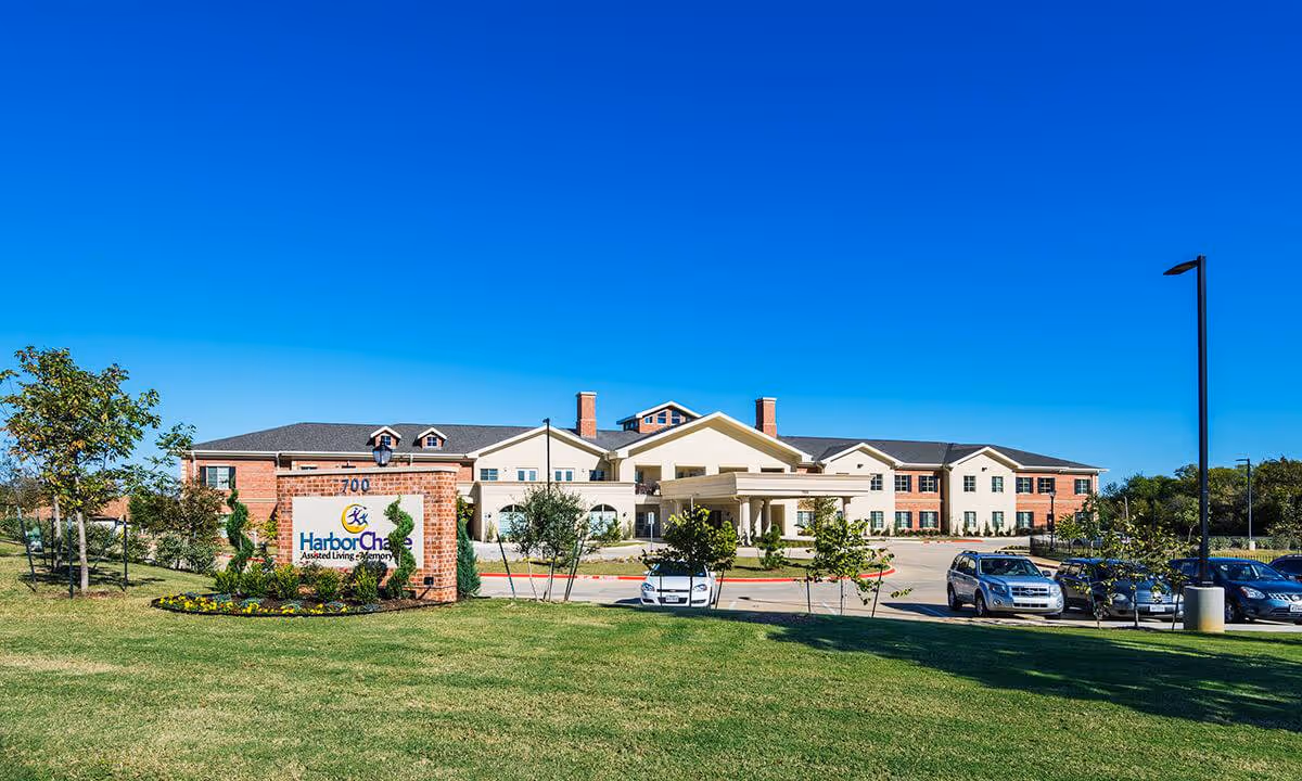 Front exterior of the HarborChase assisted living facility with a landscaped lawn, entrance drive and parked cars under a clear blue sky.