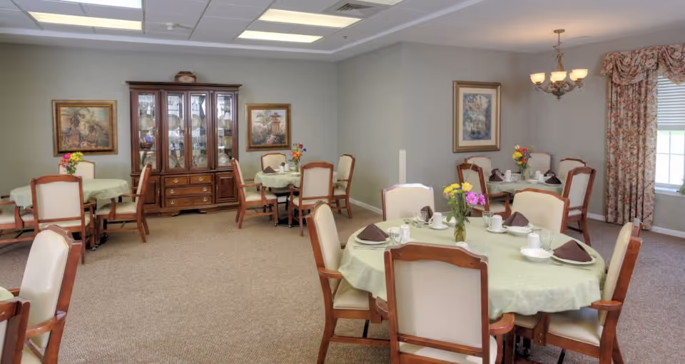 A dining room with multiple round tables covered with light green tablecloths. Each table is set with white plates, cups, glasses, and brown folded napkins. There are wooden chairs with cream-colored cushions around the tables. A wooden china cabinet with glass doors displaying dishes is against the wall, flanked by two framed paintings. A chandelier hangs from the ceiling near a window with floral curtains.