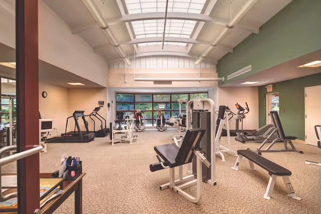 A spacious fitness room in a senior living facility with various exercise equipment including treadmills, stationary bikes, weight machines, and benches. The room has large windows letting in natural light and a high ceiling with skylights.