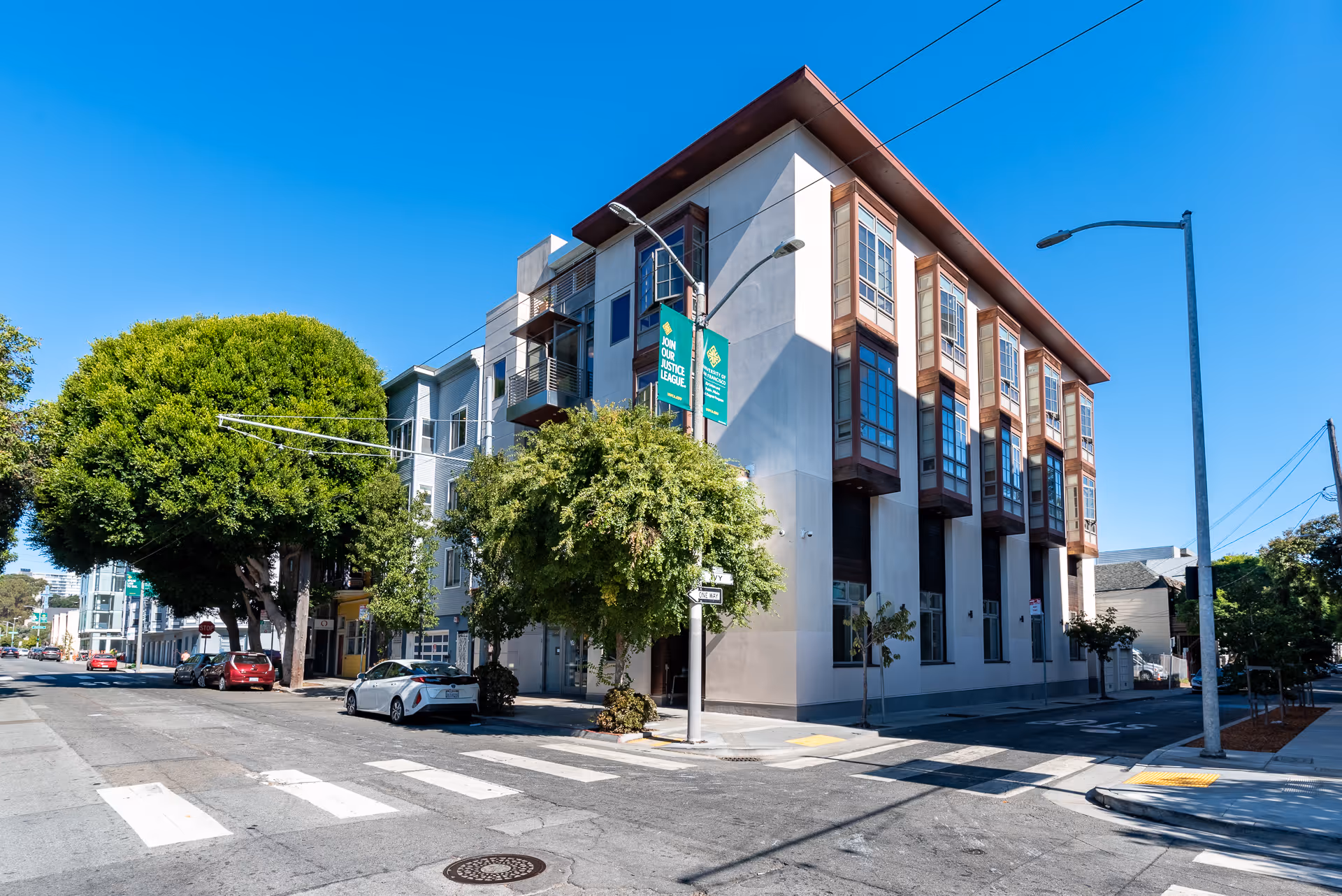 Street view of a modern multi-story building on a sunny day with clear blue sky. The building has large windows and is located at a street corner with trees and parked cars nearby.