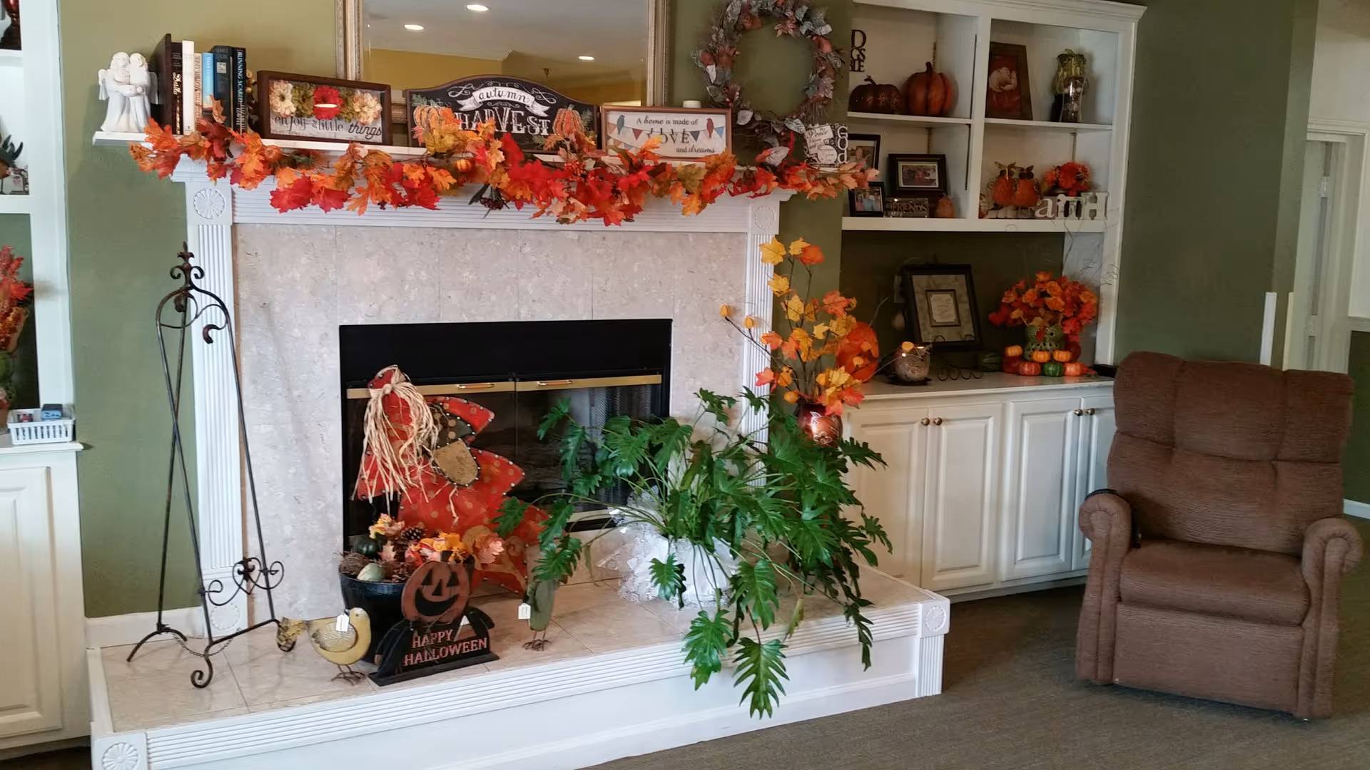 A cozy living room area featuring a white fireplace decorated with autumn and Halloween-themed items including a garland of orange and red leaves, a scarecrow figure, and a 'Happy Halloween' sign. To the right of the fireplace is a built-in white cabinet with shelves displaying seasonal decorations such as pumpkins and flowers. A comfortable brown recliner chair is positioned next to the cabinet.