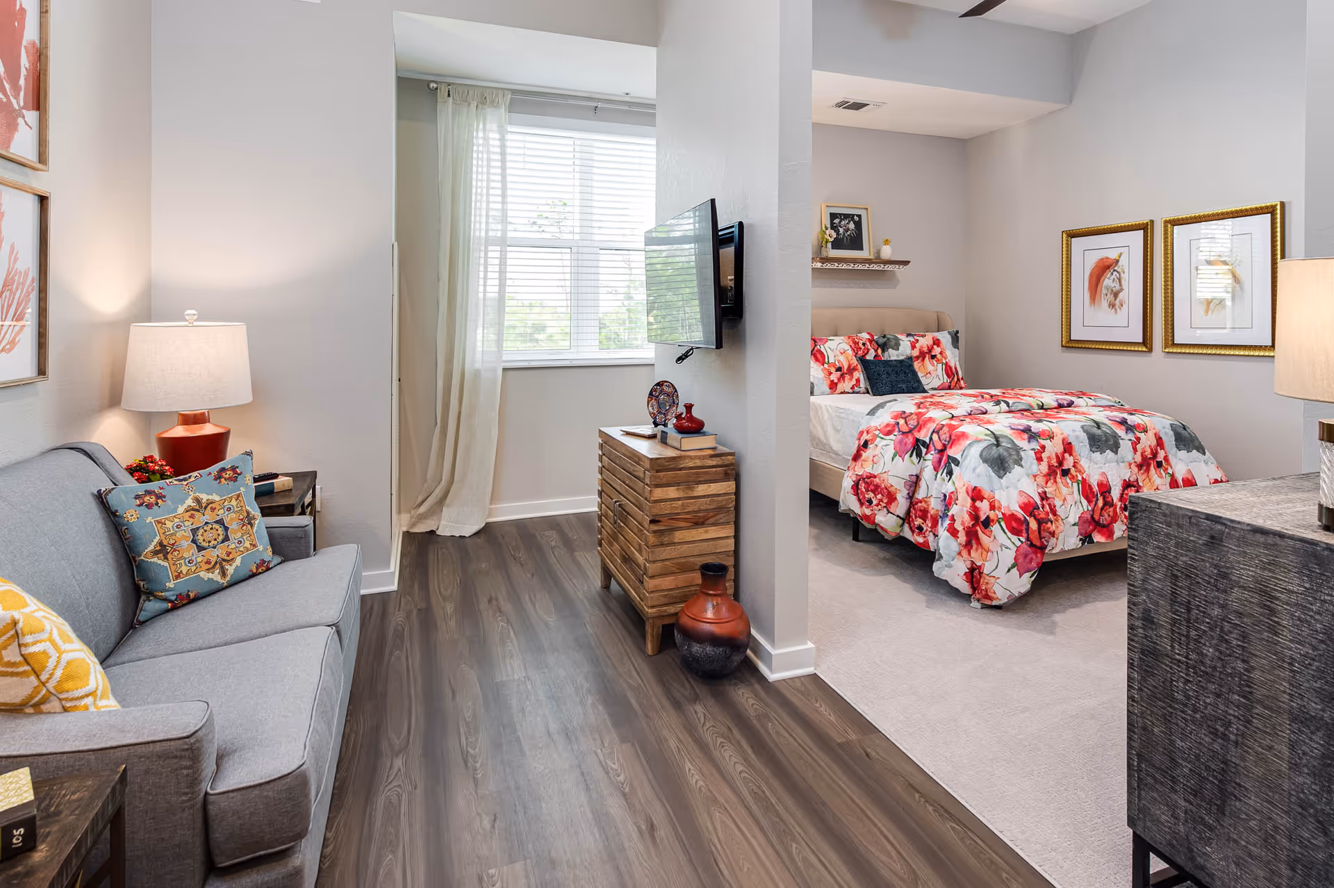 A cozy senior living space featuring a small living room with a gray sofa adorned with decorative pillows, a side table with a lamp, and framed artwork on the wall. Adjacent to the living room is a bedroom area with a bed covered in a floral comforter, two framed pictures above the bed, a nightstand with a lamp, and a wooden dresser with decorative items. The room has wood flooring in the living area and carpet in the bedroom, with a window letting in natural light.