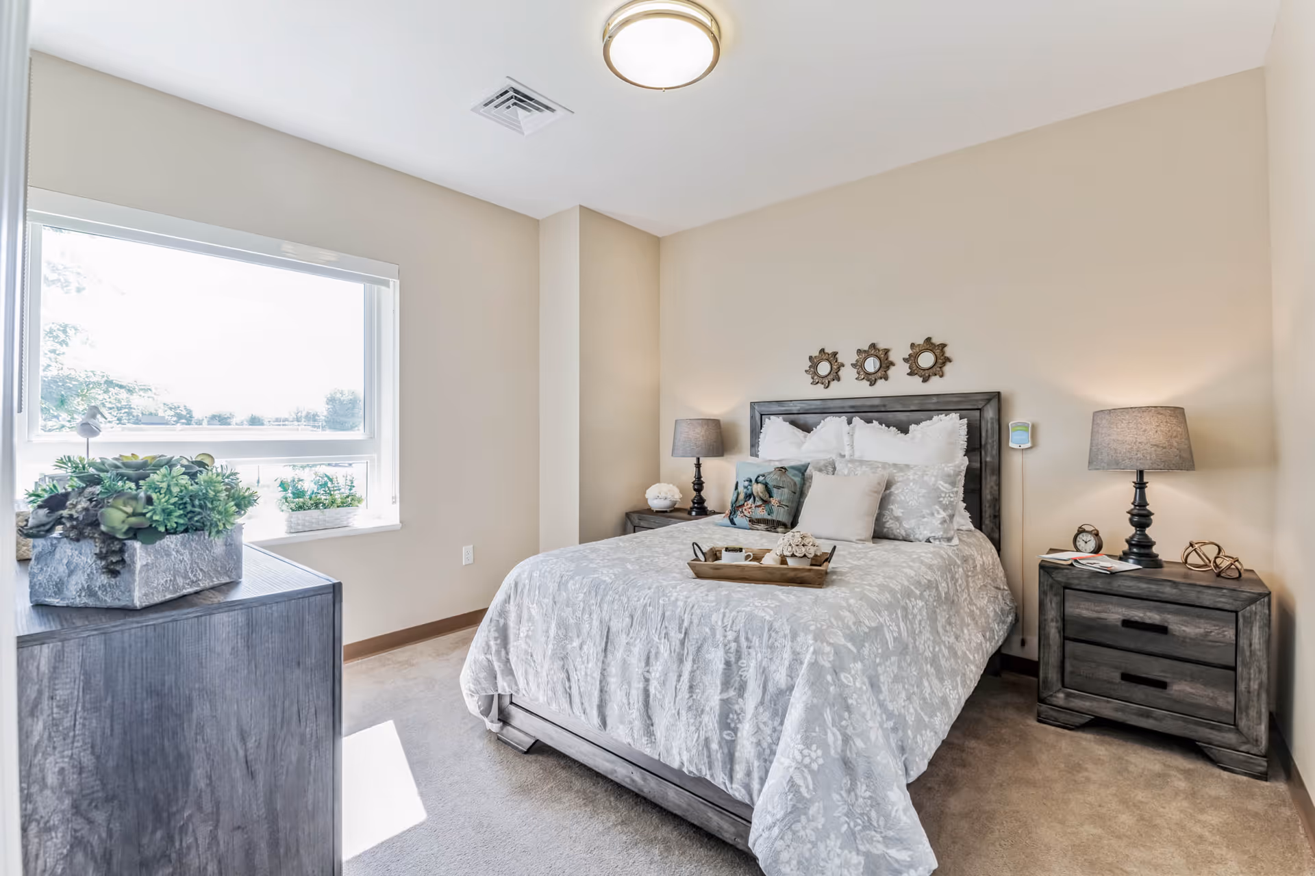 A bright and cozy bedroom with a large window letting in natural light. The room features a queen-sized bed with a gray floral bedspread, multiple pillows, and a decorative tray with a cup and flowers. There are two matching dark wood nightstands with lamps on either side of the bed. A dresser with a plant on top is positioned near the window. The walls are painted beige and three small decorative mirrors hang above the bed.