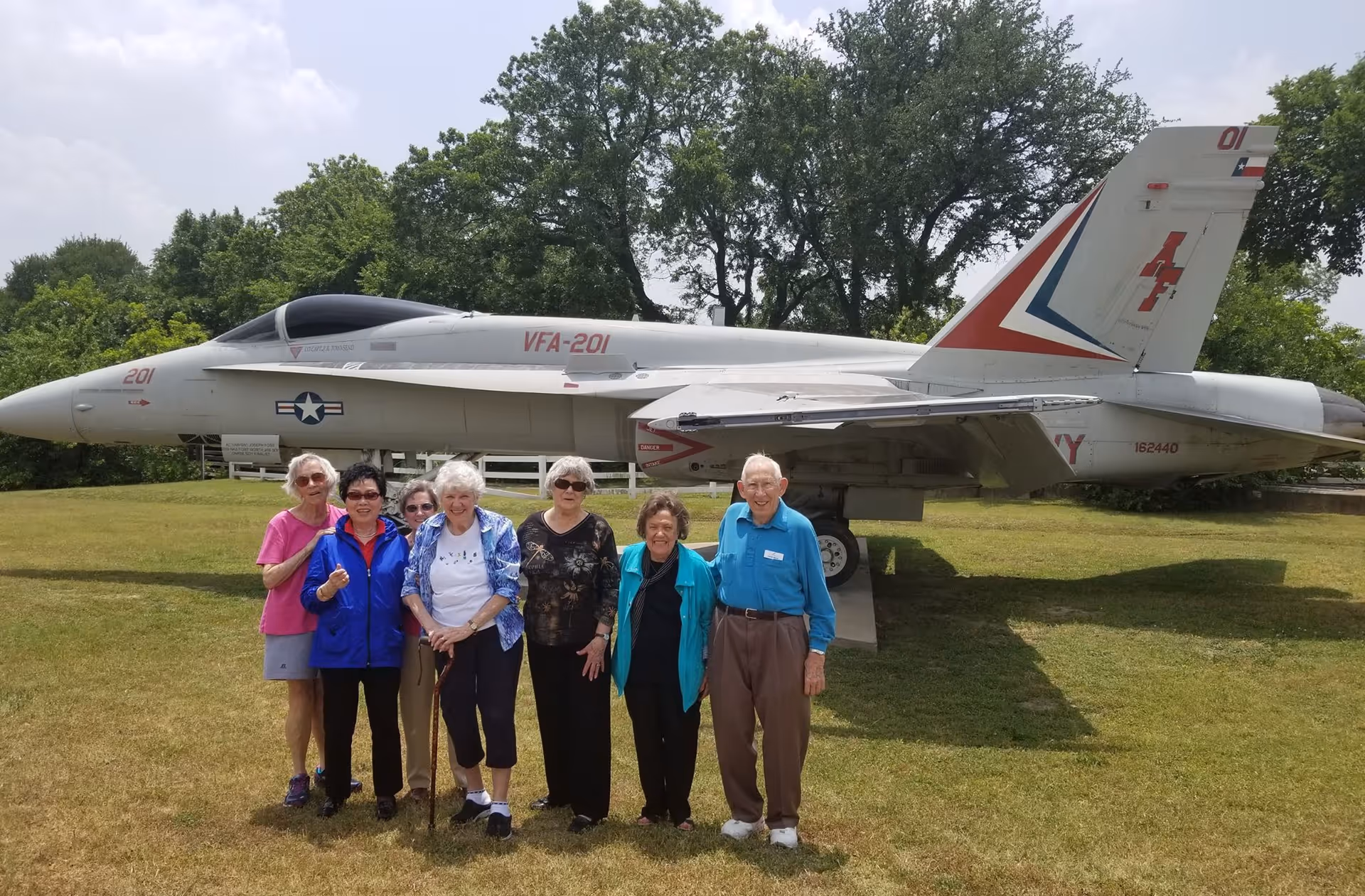 A group of seven elderly people standing on grass in front of a military jet aircraft displayed outdoors, with trees in the background and a partly cloudy sky.