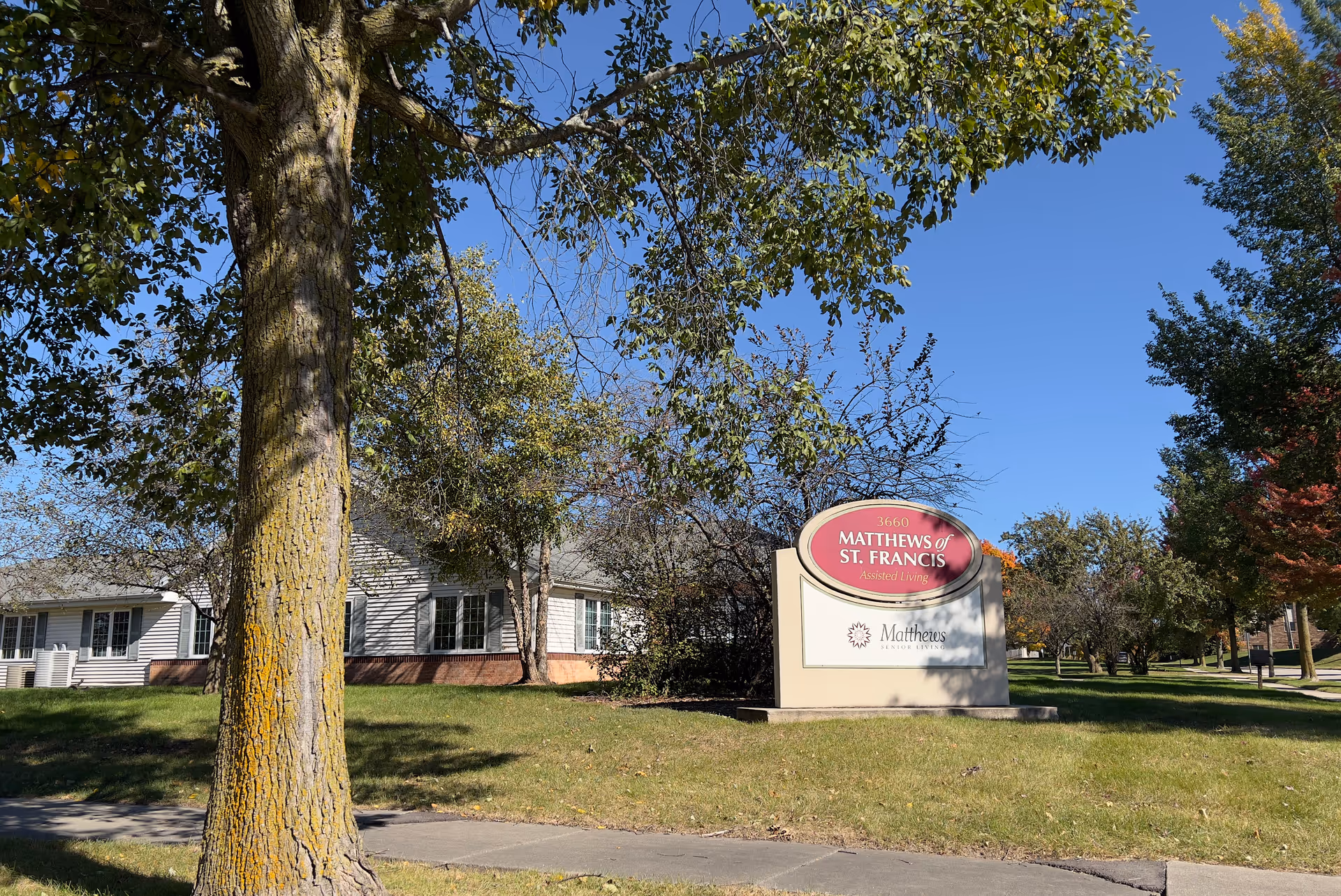Large tree and grassy lawn in front of a sign for Matthews of St. Francis Assisted Living with a single-story building and clear blue sky.