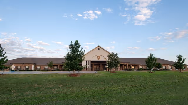 Wide exterior view of Kemp Care Center building with a large green lawn in the foreground, several small trees, and a partly cloudy sky.
