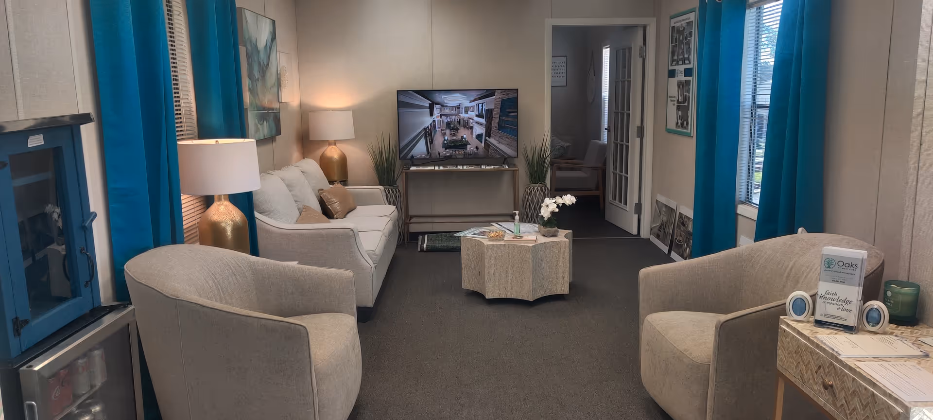 A cozy living room with two beige armchairs and a beige sofa arranged around a small hexagonal coffee table. A flat-screen TV is mounted on a stand against the far wall, with a lamp and decorative plants on either side. Blue curtains frame the windows, and there is a small table with informational brochures on the right side of the room. The room has a carpeted floor and neutral-colored walls.
