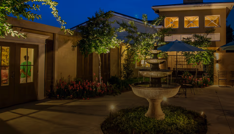 Evening view of an outdoor courtyard at Fellowship Brookside Assisted Living Community featuring a three-tier stone fountain surrounded by greenery, patio tables with umbrellas, and a building with lit windows in the background.