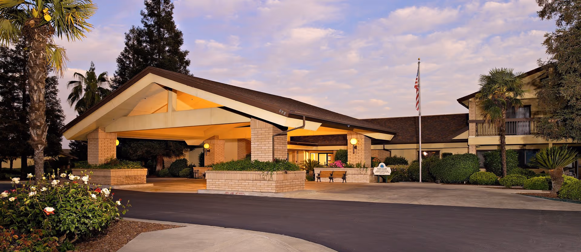 Exterior view of Palm Village Retirement Community entrance at dusk, featuring a covered drop-off area with brick pillars, landscaped bushes and flowers, palm trees, and an American flag on a flagpole.