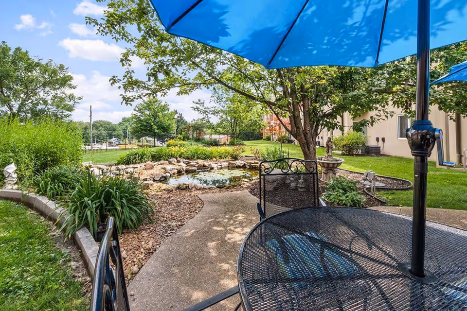 Outdoor patio area with a black metal table and chairs under a blue umbrella, surrounded by greenery, trees, and a small pond with rocks. A building is visible in the background under a partly cloudy sky.