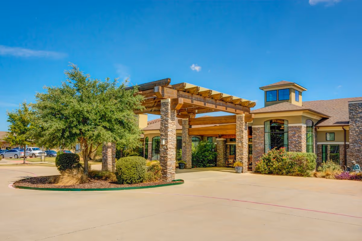 Front entrance of a senior living facility with a wooden pergola/porte-cochere supported by stone columns, landscaped shrubs, and a clear blue sky.