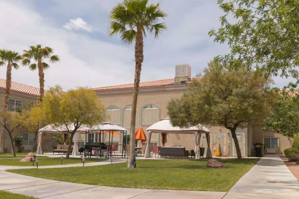 Outdoor area of Desert View Senior Living featuring green grass, palm trees, and other trees. There are two white canopy tents with outdoor seating and tables underneath. A building with beige walls and red-tiled roof is visible in the background under a partly cloudy sky.