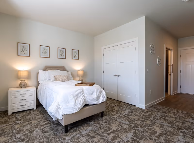 Bright, neutrally decorated bedroom with a made bed topped by a tray, matching nightstands and lamps, framed botanical prints above the headboard, and double closet doors.