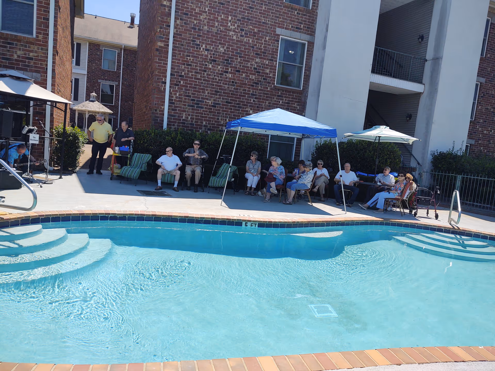 A group of elderly people sitting and relaxing by a swimming pool at an outdoor area of a senior living community. Some are seated under a blue canopy and an umbrella, with brick buildings in the background.